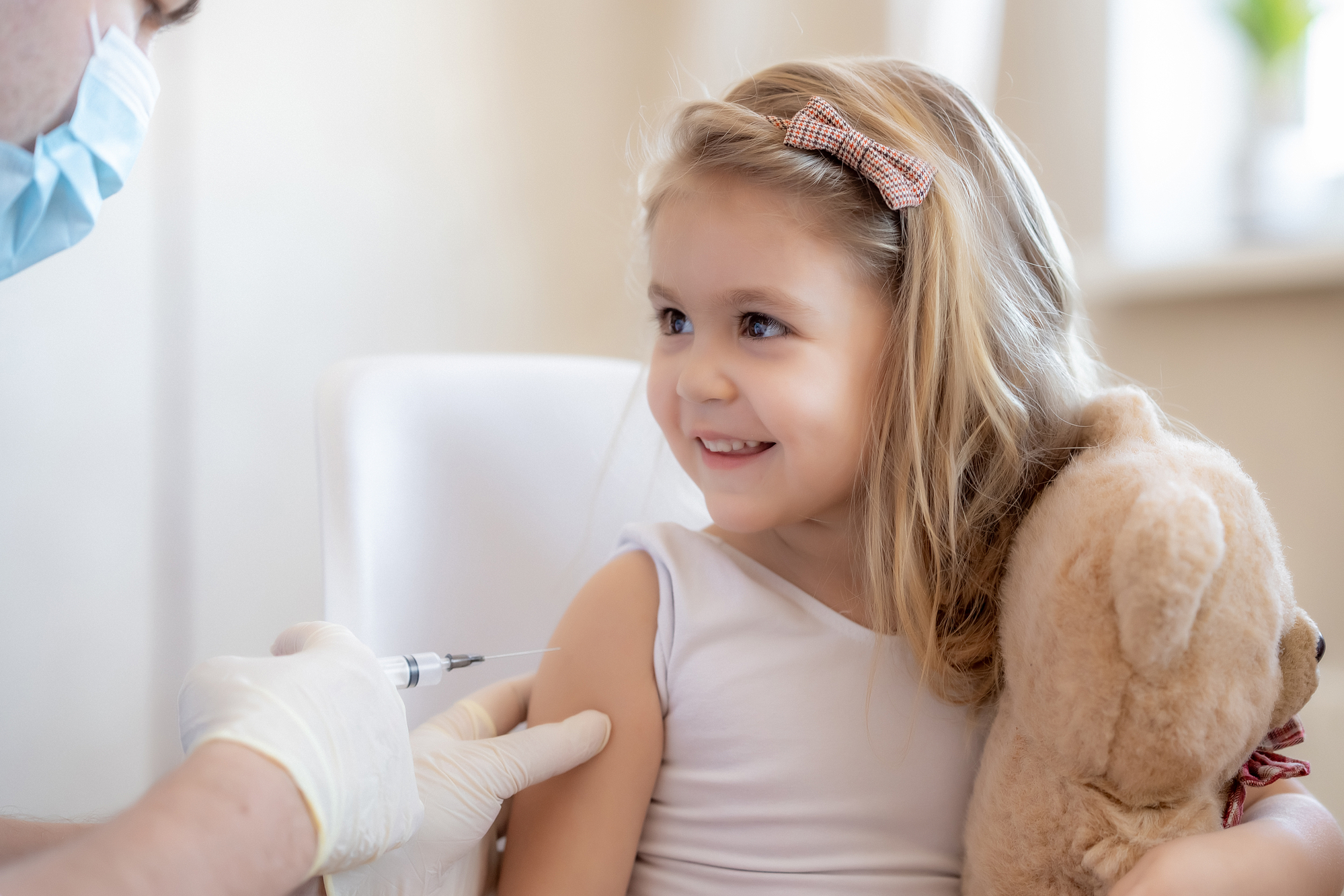 A smiling young girl receiving a flu vaccine in her arm while holding a teddy bear
