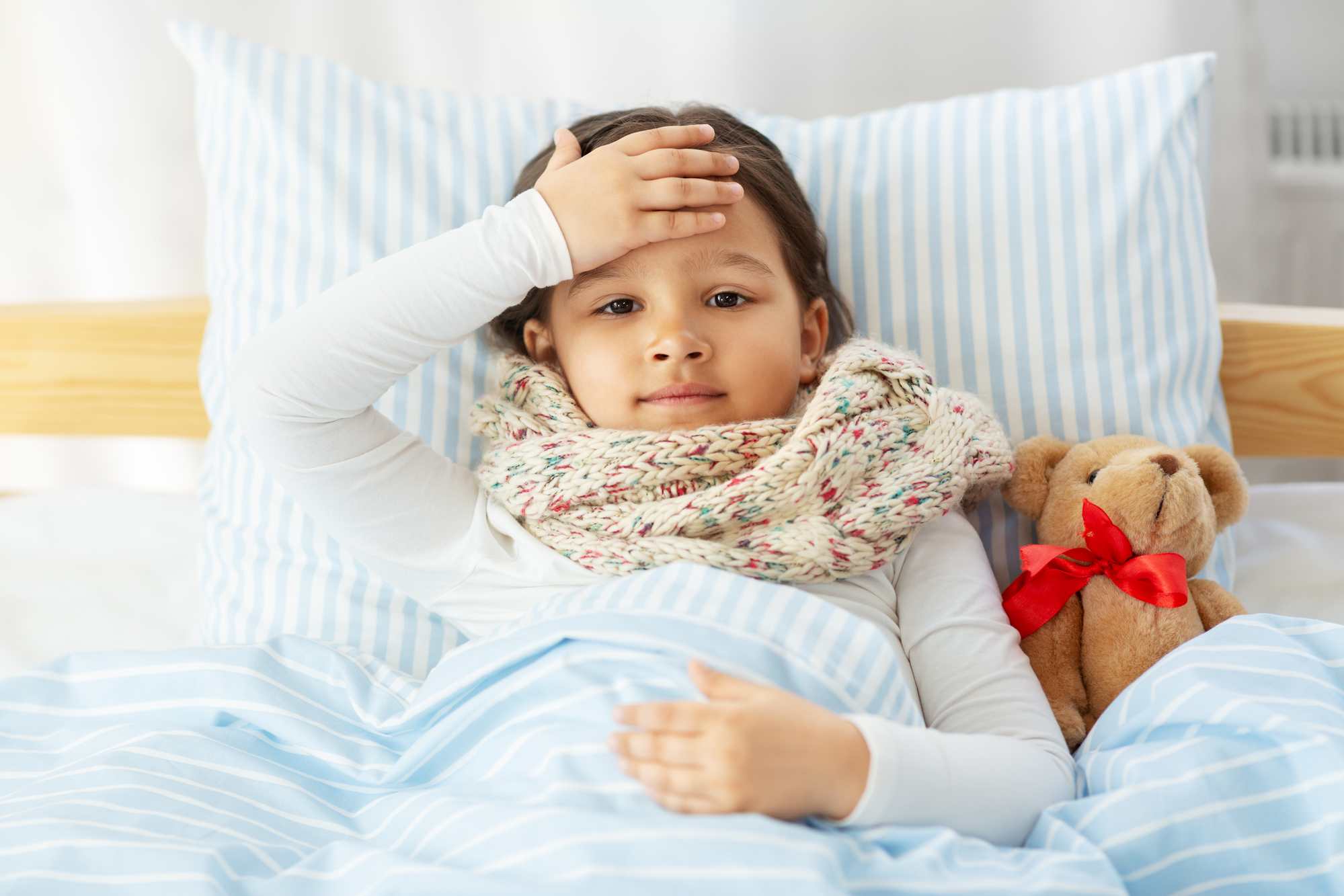 A young child lies in bed with a thermometer in their mouth, looking unwell, accompanied by a teddy bear with a red bow