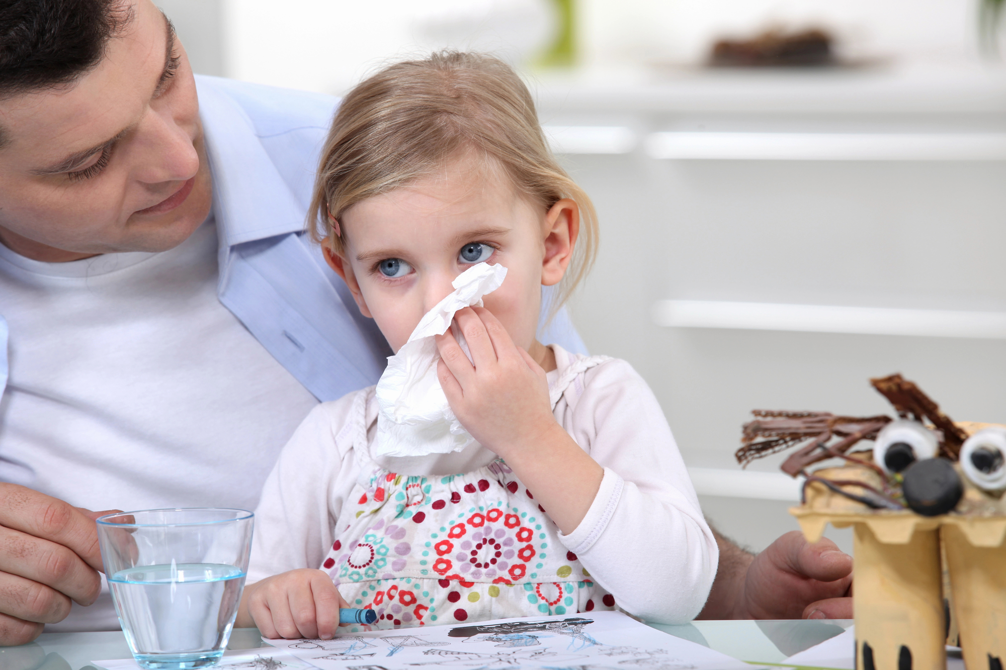 A concerned father sits beside his young daughter who is wiping her nose with a tissue, with a glass of water and a playful handmade owl craft on the table