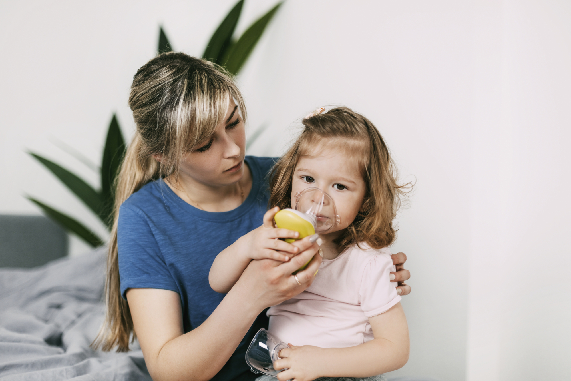 A woman in a blue shirt is attentively helping a toddler girl drink from a small glass cup.