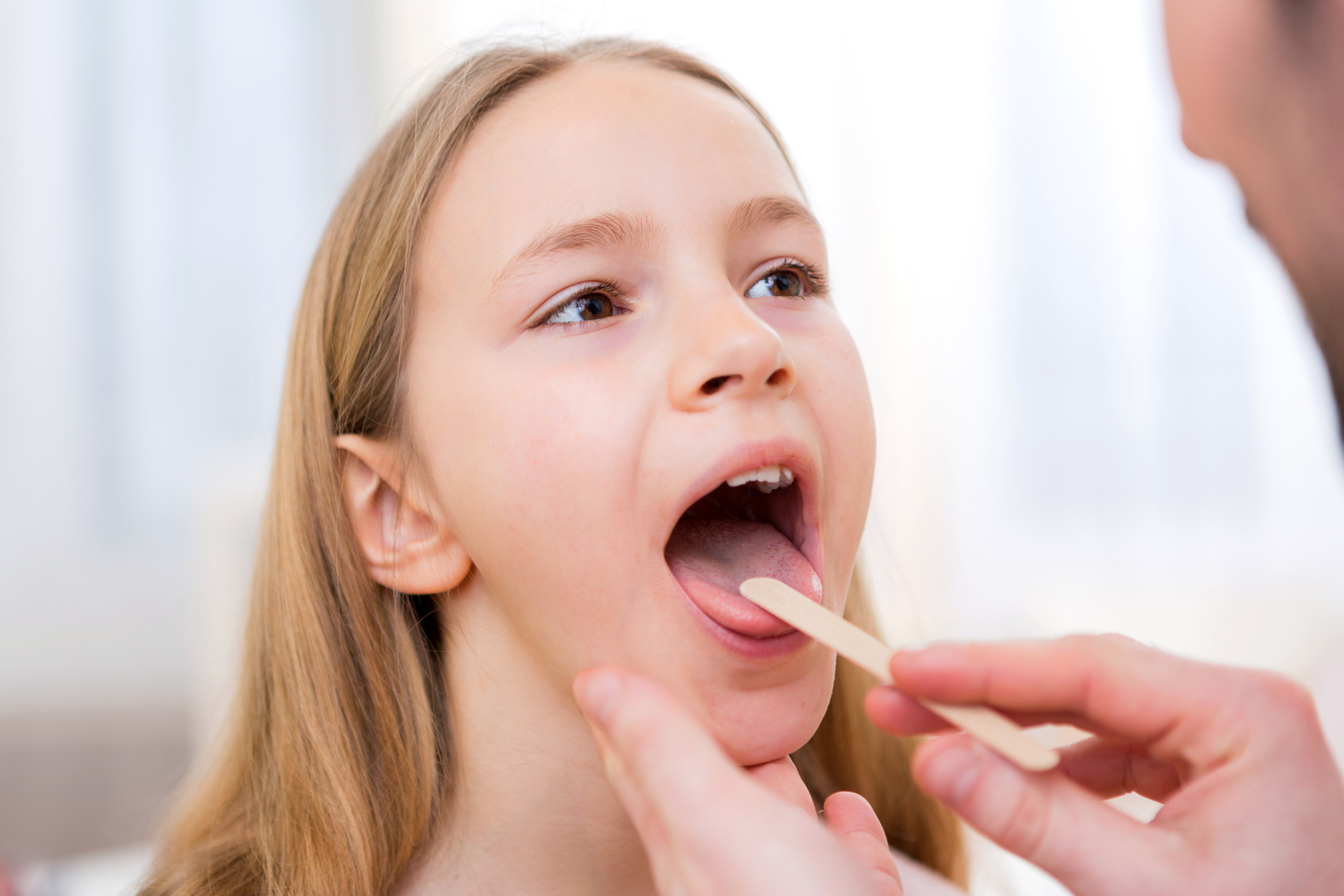 A young girl opens her mouth wide for a throat examination by a healthcare professional.